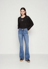 Gilet en dentelle noire sur une chemise noire ajustée avec motif soleil, associé à un jean évasé bleu clair. Des chaussures décontractées complètent le look.