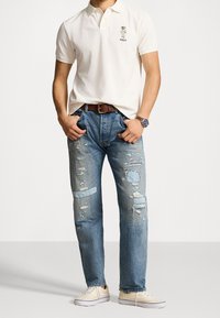 Beige pikétröja med broderad hundlogotyp, kombinerad med slitna blå jeans med lappar och beige sneaker. Klockan på handleden kompletterar den avslappnade stilen.