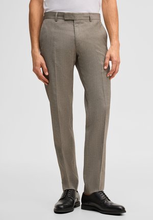 Mann trägt beige, gerade geschnittene Anzughose mit schwarzen Leder-Schnürschuhen vor einfarbigem Hintergrund.