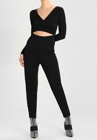 Mono negro de mangas largas y diseño cruzado, con un corte en el abdomen. Combinado con botas de tobillo brillantes y una silueta elegante.