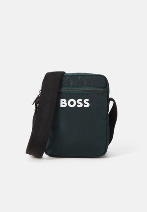 Pequeña bolsa de hombro verde oscuro con bolsillo frontal con cremallera y correa negra ancha, con la palabra "BOSS" en letras blancas.