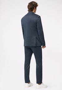 Navyblauer maßgeschneiderter Anzug aus strukturiertem Stoff, mit einer Jacke mit Schlitz und schmal geschnittenen Hosen, kombiniert mit weißen Sneakers.
