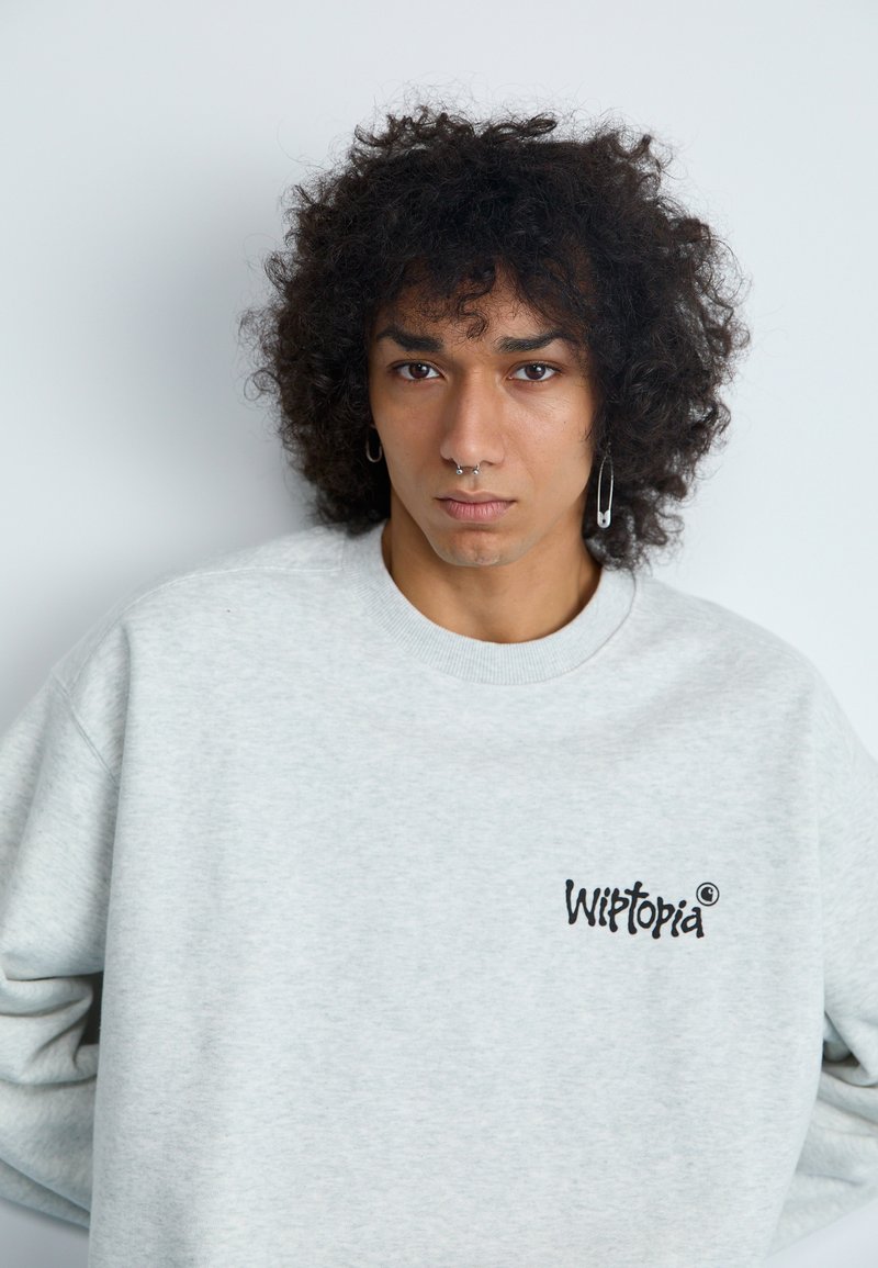 Grå sweatshirt med rundad halsringning, som har en liten svart "Witopia"-logotyp på vänster bröstområde. Mjuk, texturerad tyg.