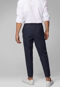 Homme portant une chemise blanche à manches longues, un pantalon noir slim ajusté et des baskets blanches, se tenant devant un fond gris uni.