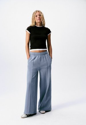 Woman with wavy blonde hair stands wearing a black cropped t-shirt with white trim, loose light blue pants, and white shoes on a white background.