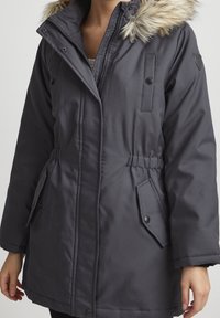 Parka grise à capuche avec bord en fausse fourrure et fermeture à pressions. Présente une taille élastique, deux poches latérales et un design structuré.
