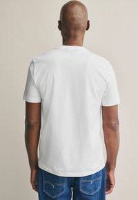 Weißes, kurzärmliges T-Shirt mit Rundhalsausschnitt, aus weichem Baumwollmaterial, kombiniert mit blauen Jeans aus Denim, die eine Rückentasche mit Stichdetails aufweisen.