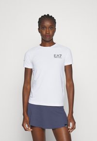 Witte korte mouw atletische shirt met EA7-logo op de linker borst. De stof lijkt lichtgewicht en ademend. Gecombineerd met een marineblauwe geprinte rok.