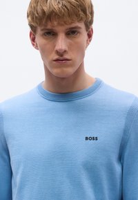 Világoskék kötött anyagból készült kerek nyakú pulóver, bordázott nyakrésszel és egy kis fekete "BOSS" logóval a mellkason.