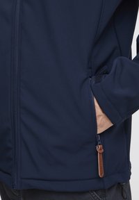 Veste softshell marine avec une texture lisse, fermeture zippée intégrale, poches latérales et détail de tirette en cuir.
