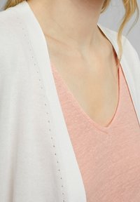 Valkoinen neuletakki, jossa on huomaamaton sivupisto, päällystetty pehmeällä vaaleanpunaisella V-pääntiellä. Molemmissa vaatteissa on kevyttä, tekstuurista kangasta.