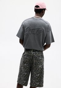 Grå bomulds T-shirt med tekstureret logo på ryggen, parret med knælange leopardprint shorts. Accessoriseret med et sort bælte og en lyserød kasket.
