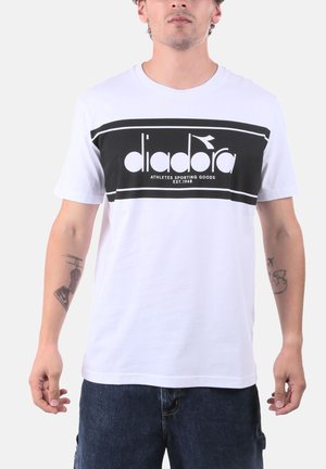 Uomo che indossa una T-shirt bianca con il logo rettangolare nero Diadora e jeans blu scuro, in piedi davanti a uno sfondo semplice.