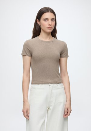 Donna con capelli lunghi e scuri che indossa una maglietta beige a maniche corte e pantaloni bianchi a vita alta, in piedi davanti a uno sfondo bianco semplice.