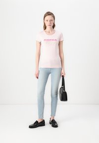 T-shirt ajustée rose avec un graphique "GUESS LOS ANGELES" en dégradé de couleurs, associée à un jean skinny bleu clair et des mocassins noirs, tenant un sac noir.