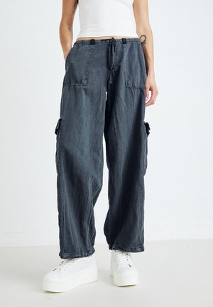 Persona che indossa pantaloni cargo scuri e larghi con grandi tasche davanti e ai lati, sneakers bianche con plateau e un top corto bianco.