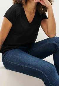T-shirt en coton noir avec un col en V et des manches courtes roulées, associé à un jean en denim bleu foncé. Le tissu semble lisse et légèrement texturé.