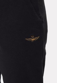 Pantaloni sportivi neri con una trama morbida. Dettagli notevoli includono un emblema piumato ricamato in oro sulla tasca frontale.
