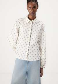 Vit skjorta med en vågig krage, svart blommönster och långa puffärmar; matchad med en ljus denimkjol.