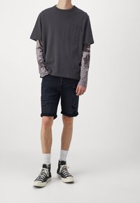 Camiseta gris de manga corta con bolsillo, superpuesta sobre una camiseta de manga larga con estampado, combinada con pantalones cortos vaqueros oscuros y zapatillas negras de caña alta.