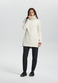 Witte softshell jacket met een hoge kraag, ritssluiting zijzakken en contrasterende zwarte accenten. Gdragend met zwarte broek en sneakers.