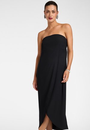 Vrouw die een strapless zwarte midi-jurk draagt met overslagrok, zilveren armband en grote statement oorbellen, staand tegen een effen witte achtergrond.