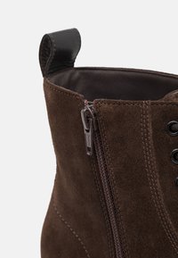 Brown suede ankle boot featuring a zipper closure, round laces, and a stitched design. Includes a leather pull tab at the top.