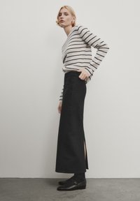Jupe maxi en denim noir avec fente latérale, associée à un pull rayé crème et noir. Le modèle porte des chaussures plates noires. Design simple et moderne.