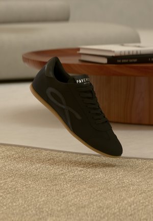 Zwarte sneaker met een gumzool en logo aan de zijkant, zwevend boven een beige tapijt voor een houten salontafel met gestapelde boeken.