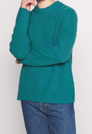 Mann trägt einen türkisfarbenen Strickpullover und blaue Jeans, steht mit einem Arm verschränkt und der Hand am Kinn vor einem einfarbigen Hintergrund.
