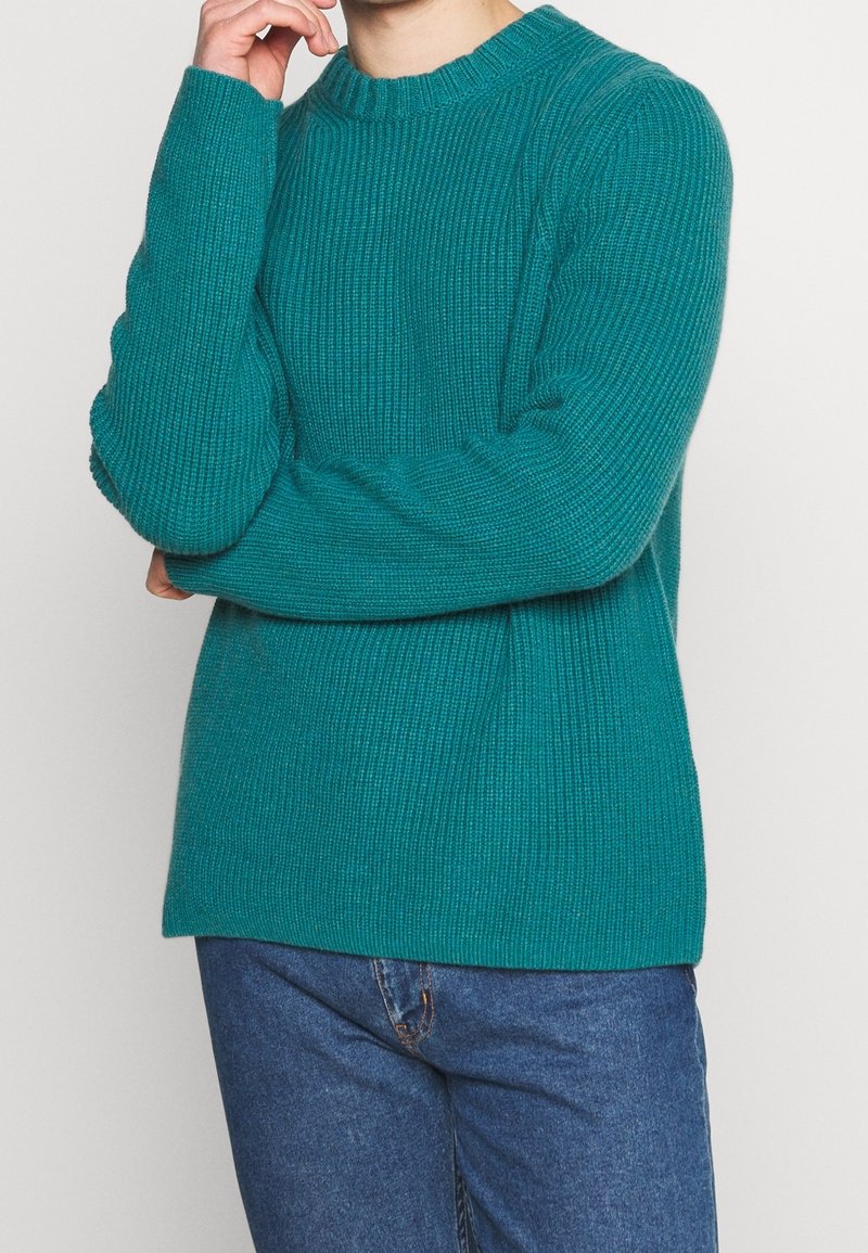 Homme portant un pull en tricot bleu sarcelle et un jean bleu, avec un bras croisé et la main touchant le menton, se tenant devant un fond uni.