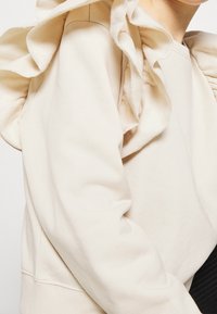 Ljusbeige sweatshirt med volangdetaljer på axlarna, med en mjuk textur och avslappnad passform. Ribbad mudd och fåll.