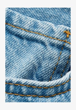 Niebieska tkanina dżinsowa o teksturowanym splocie, z kontrastującymi żółtymi przeszyciami wzdłuż szwów i fałd. Zbliżenie na szczegóły materiału.