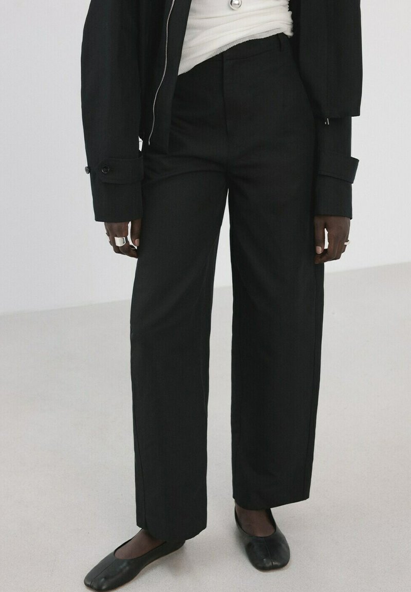 Personne portant un pantalon large noir taille haute, une veste noire, un haut blanc, des chaussures plates noires et des bagues argentées aux doigts, debout sur un sol clair.