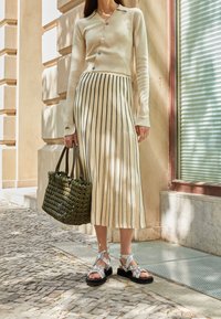 Cárdigan beige claro con botones, falda midi a rayas, bolso de mimbre verde y sandalias plateadas con tiras y suelas negras.