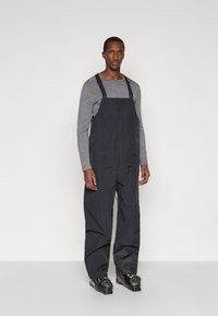 Άντρας που φοράει μαύρα παντελόνια σνόουμπουτ, γκρι μπλουζάκι με μακριά μανίκια και μαύρες μπότες σκι, στέκεται μπροστά σε λευκό φόντο.