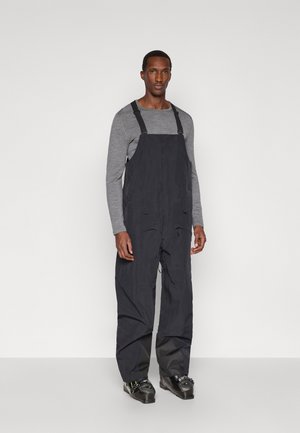 Homem em calças de neve pretas, camisa de manga longa cinza e botas de esqui pretas, de pé contra um fundo branco liso.