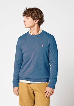 Jeune homme aux cheveux bouclés portant un pull bleu sur une chemise beige et un short moutarde, regardant vers sa gauche sur un fond blanc.