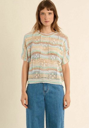 Giovane donna che indossa una maglietta corta a maniche corte larga, multicolore e a maglia aperta, e jeans blu a vita alta su uno sfondo semplice.