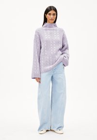 Fliederfarbener Strickpullover mit hohem Kragen und weiten Ärmeln, kombiniert mit hellblauen, weit geschnittenen Jeans und weißen Schuhen. Strukturiertes Kabelstrickdesign.