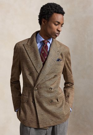 Homme portant un blazer croisé à carreaux marron, une chemise bleue, une cravate rouge à motifs et un pantalon gris, regardant vers le bas les mains dans les poches.