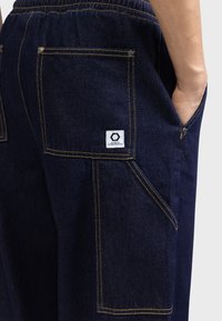 Dunkelblaue Jeans mit elastischem Bund, zwei Gesäßtaschen, gelben Nähten und einem kleinen weißen Etikett mit Logo.