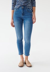 Jeans de mezclilla azul con un corte ajustado, cintura de altura media, diseño de cinco bolsillos y un ligero desgaste. Combinados con mocasines de color claro.