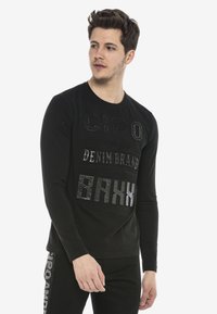 Schwarzes Langarmshirt mit texturiertem metallischem Schriftzug und Markengrafiken in Grau. Einfacher Rundhals und taillierte Passform.