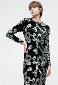 Mulher vestindo um vestido longo de veludo preto com padrões brilhantes de flores e ramos prateados, posando com uma mão na anca.