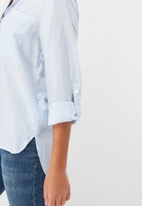Camicia a righe blu chiaro con taschino sul petto, maniche arrotolate e orlo curvo. Realizzata in tessuto leggero con una consistenza fresca.