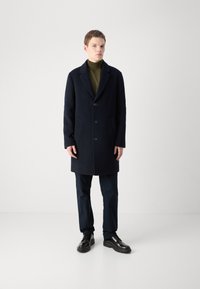 Marineblauer Wollmantel mit eingeschnittenen Revers, zwei frontalen Taschen und drei Knöpfen. Kombiniert mit einem dunkelgrünen Rollkragenpullover und navyfarbener Hose.