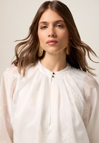Blusa blanca con delicados bordados, escote redondo, detalles de botones y mangas largas. La modelo lleva pendientes dorados con textura.