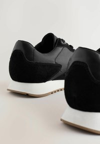 Ein Paar schwarze Sportsneaker mit Wildleder- und Stoffdetails, weißen Zwischensohlen und hellbrauner Außensohle, auf einer hellen Oberfläche platziert.