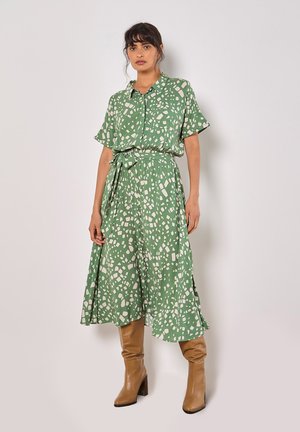 Grünes Kleid mit Hemdkragen, kurzen Ärmeln und einem Bindegürtel. Hat ein Muster aus unregelmäßigen cremigen Formen; der Stoff wirkt leicht.